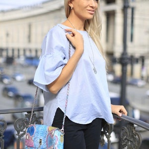 May include: A light blue top with ruffled sleeves and a patterned handbag with a chain strap. The handbag features a tassel and a mix of blue, pink, and yellow colors. The person is wearing black pants and a silver necklace.
