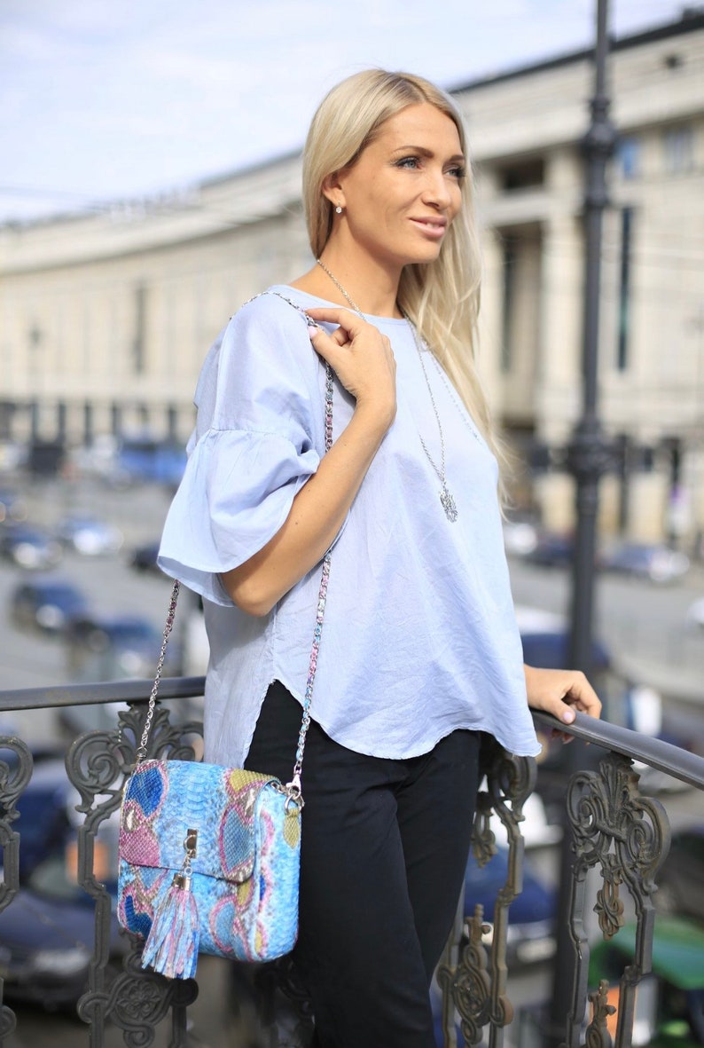 May include: A light blue top with ruffled sleeves and a patterned handbag with a chain strap. The handbag features a tassel and a mix of blue, pink, and yellow colors. The person is wearing black pants and a silver necklace.