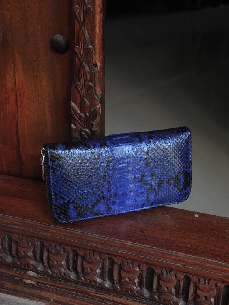 Op de afbeelding: Een blauwe en zwarte slangenleren portemonnee met een ritssluiting. De portemonnee ligt op een houten oppervlak met ingewikkelde houtsnijwerk.