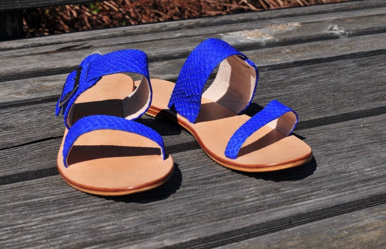 May include: A pair of blue sandals with a textured, snakeskin-like pattern. The sandals have a light brown sole and straps with a buckle closure. The sandals are displayed on a weathered wooden surface.