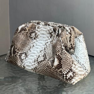 May include: A clutch bag with a snakeskin pattern in shades of white, brown, and tan. The bag has a gathered top and a rectangular shape. The snakeskin pattern is visible across the entire surface of the bag.