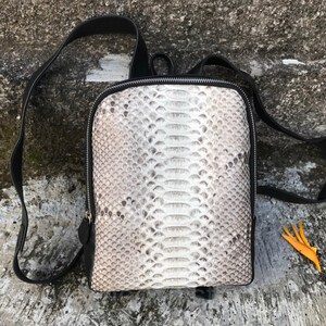 以下が含まれることがあります： 白とグレーのヘビ柄の小さな黒い革のバックパック。バックパックはジッパーで閉じられ、調節可能なストラップが付いています。