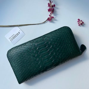 以下が含まれることがあります： ジッパー留めのダークグリーンのヘビ革財布。財布には「#RECOONIST 1  ITEM NO. 0200150」というテキストが書かれたタグが付いています。