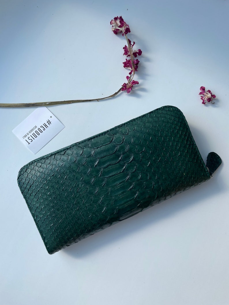 以下が含まれることがあります： ジッパー留めのダークグリーンのヘビ革財布。財布には「#RECOONIST 1  ITEM NO. 0200150」というテキストが書かれたタグが付いています。