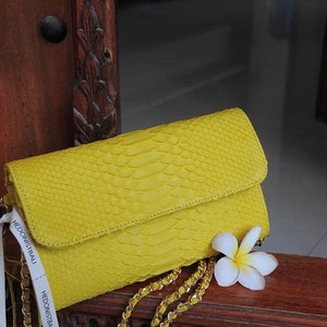 May include: A bright yellow snakeskin clutch purse with a gold chain strap and a white flower. The purse has a tag that reads "Hedonist Bali".