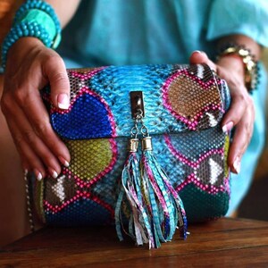 May include: A colorful, patterned handbag with a flap closure and a decorative tassel. The bag features a mix of blue, pink, green, and brown hues, resembling a snakeskin design. The bag is held by a person.
