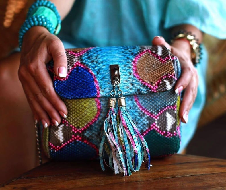 May include: A colorful, patterned handbag with a flap closure and a decorative tassel. The bag features a mix of blue, pink, green, and brown hues, resembling a snakeskin design. The bag is held by a person.