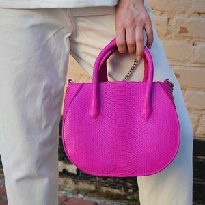 Puede incluir: Un bolso de mano de piel de serpiente de color rosa brillante con una correa de cadena plateada. El bolso tiene una forma redondeada y dos asas superiores.