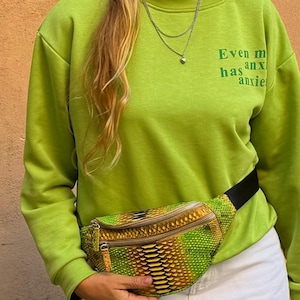 Op de afbeelding: Een limoengroen sweatshirt met de tekst "Even my anxiety has anxiety" erop gedrukt. De persoon draagt ​​een heuptas met een slangenhuidpatroon en een zwarte band.