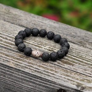 Puede incluir: Un brazalete de piedra de lava negra con un dije de Buda plateado. El brazalete está sobre una superficie de madera.