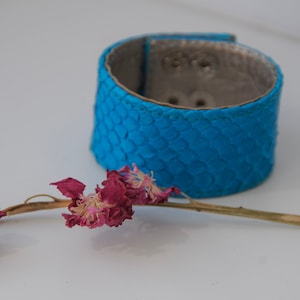 Könnte beinhalten: Ein blaues Lederarmband mit schuppiger Textur. Im Vordergrund befindet sich eine getrocknete rosa Blume.