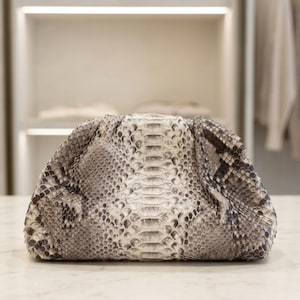 May include: A clutch handbag featuring a textured snakeskin pattern in shades of grey, black, and white. The bag has a rounded, gathered shape with a central vertical panel of scales and a soft, slouchy silhouette without visible hardware or straps.