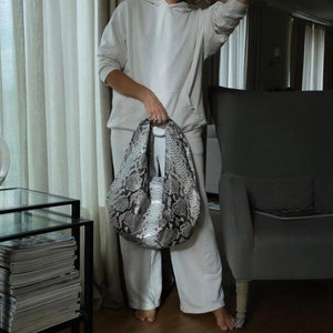 May include: A woman wearing a white hoodie and matching sweatpants holds a snakeskin handbag. The bag has a light gray and black pattern. The woman is standing in a room with a chair and a table with magazines on it.