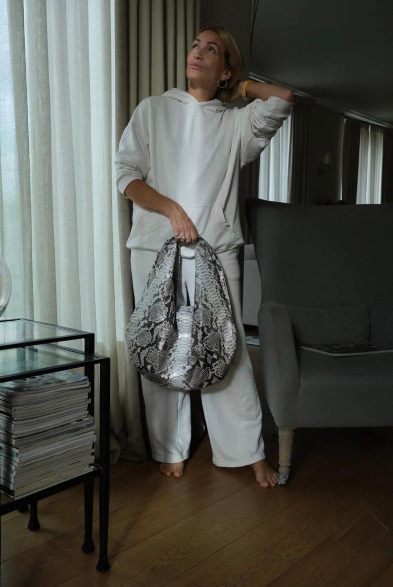 May include: A woman wearing a white hoodie and matching sweatpants holds a snakeskin handbag. The bag has a light gray and black pattern. The woman is standing in a room with a chair and a table with magazines on it.