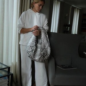 May include: A woman wearing a white hoodie and white pants is holding a large, gray and white snakeskin handbag.