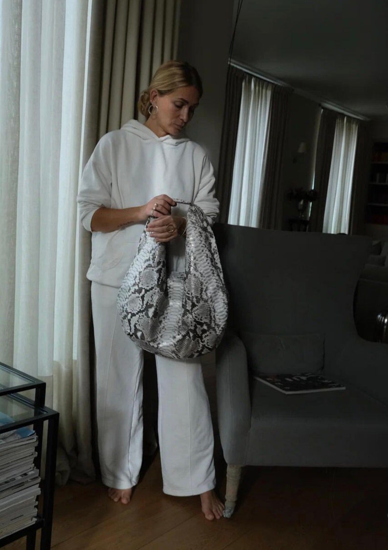 May include: A woman wearing a white hoodie and white pants is holding a large, gray and white snakeskin handbag.
