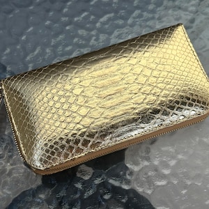 Può includere: Un portafoglio rettangolare dorato con un motivo a pelle di serpente strutturato. Ha una chiusura con cerniera e un piccolo tirazip. Il design è elegante e moderno, adatto per trasportare l'essenziale. Misura circa 20 cm x 10 cm.