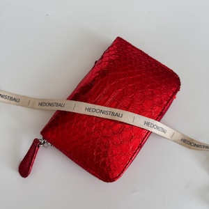 Könnte beinhalten: Eine glänzende rote Geldbörse mit strukturiertem Muster und Reißverschluss. Die Geldbörse hat einen passenden roten Zipper und ein beiges Band mit dem Aufdruck "HEDONISTBALI".