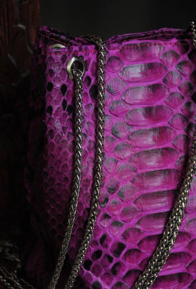 Op de afbeelding: Een close-up van een paarse slangenleren handtas met een zilveren kettingband.