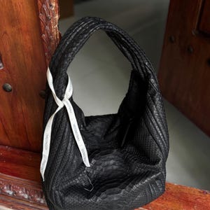 Op de afbeelding: Een zwarte handtas met een textuur, slangachtig patroon. De tas heeft een afgeronde vorm met een enkele handgreep en een wit lint met de tekst "HEDONISTBAU" aan de handgreep.