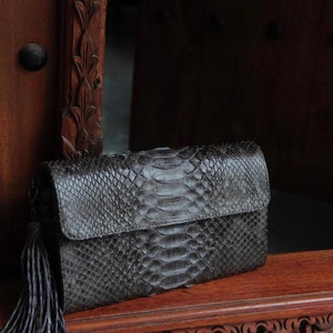 Puede incluir: Un bolso de mano negro de piel de serpiente con un borla. El bolso está sobre una superficie de madera con tallas intrincadas.