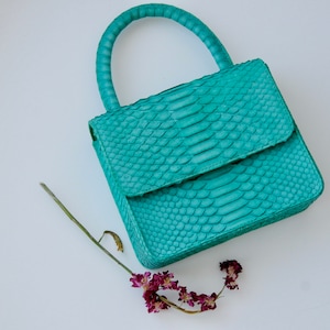 May include: A teal green snakeskin handbag with a top handle. The bag has a flap closure and a textured snakeskin pattern.