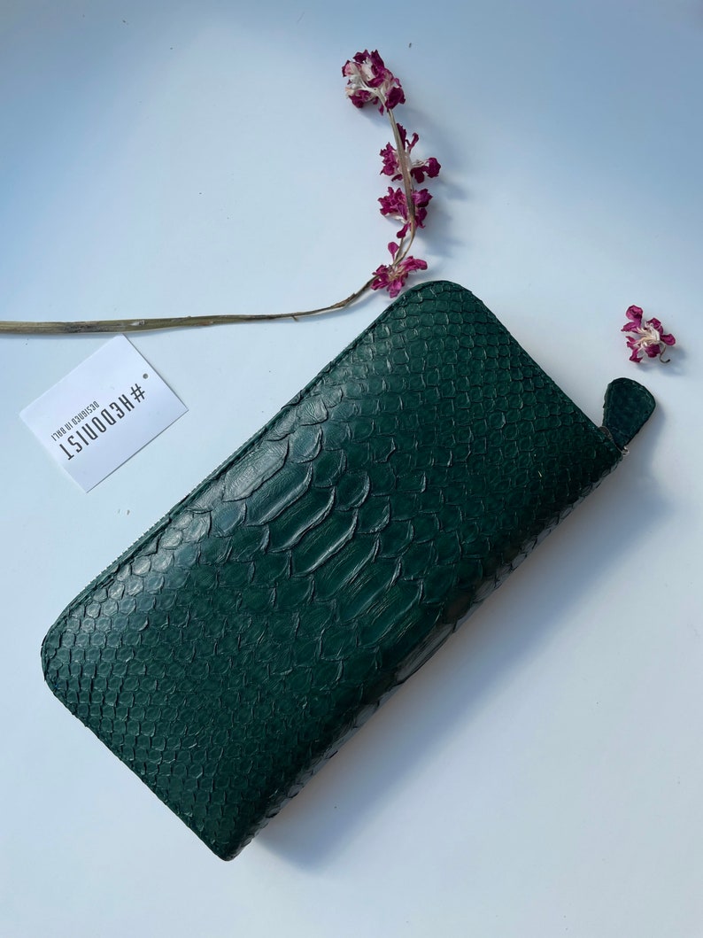 以下が含まれることがあります： ジッパー留めのダークグリーンのヘビ革財布。財布には「#RECO1151 17.99 in. 8230032」というテキストが書かれたタグが付いています。