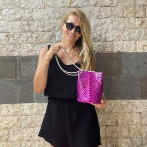 Op de afbeelding: Een vrouw in een zwarte jurk en zonnebril houdt een klein roze slangenleren handtasje vast met een zilveren kettingband.