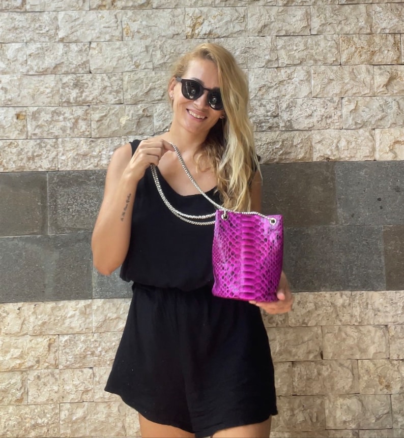 Op de afbeelding: Een vrouw in een zwarte jurk en zonnebril houdt een klein roze slangenleren handtasje vast met een zilveren kettingband.