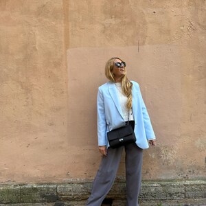 May include: A light blue blazer, white top, and gray pants ensemble. A black shoulder bag with a chain strap and white sneakers complete the outfit. The background is a textured, peach-colored wall.