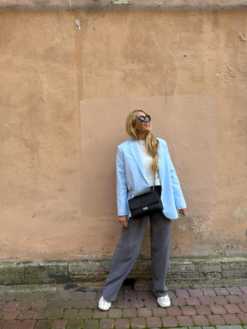 May include: A light blue blazer, white top, and gray pants ensemble. A black shoulder bag with a chain strap and white sneakers complete the outfit. The background is a textured, peach-colored wall.