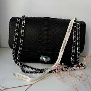 May include: Black rectangular handbag with a silver chain strap and clasp. The bag has a textured pattern and a white ribbon with the text "HEDONISTBAU". A few sprigs of pink flowers are next to the bag.