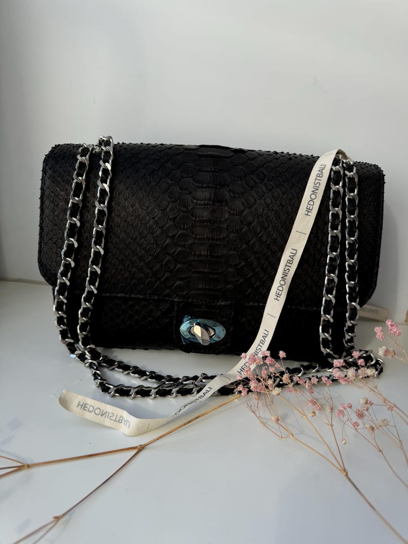 May include: Black rectangular handbag with a silver chain strap and clasp. The bag has a textured pattern and a white ribbon with the text "HEDONISTBAU". A few sprigs of pink flowers are next to the bag.