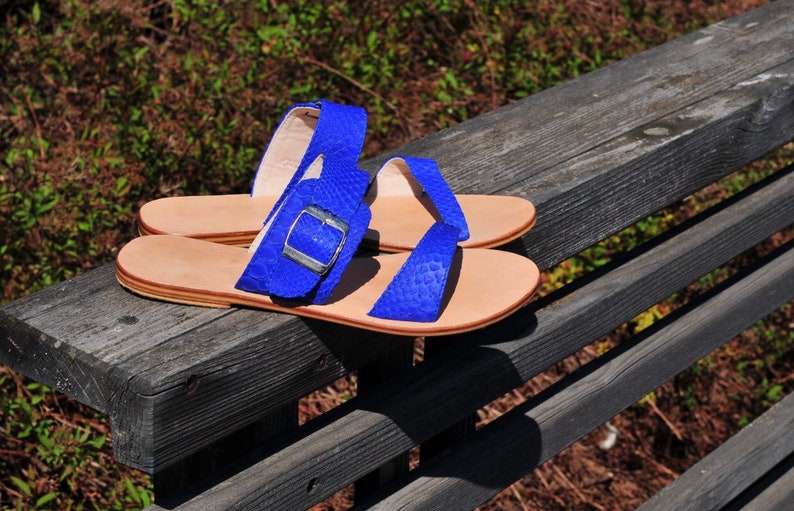 May include: A pair of blue sandals with a textured pattern, featuring a buckle closure. The sandals have light brown soles and are displayed on a weathered wooden surface. The sandals are a summer footwear.