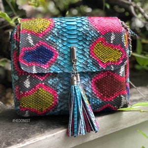 May include: A colorful crossbody bag with a snakeskin pattern in shades of blue, pink, yellow, and black. The bag features a silver chain strap and a matching tassel. The text "#HEDONIST" is visible on the bottom left.