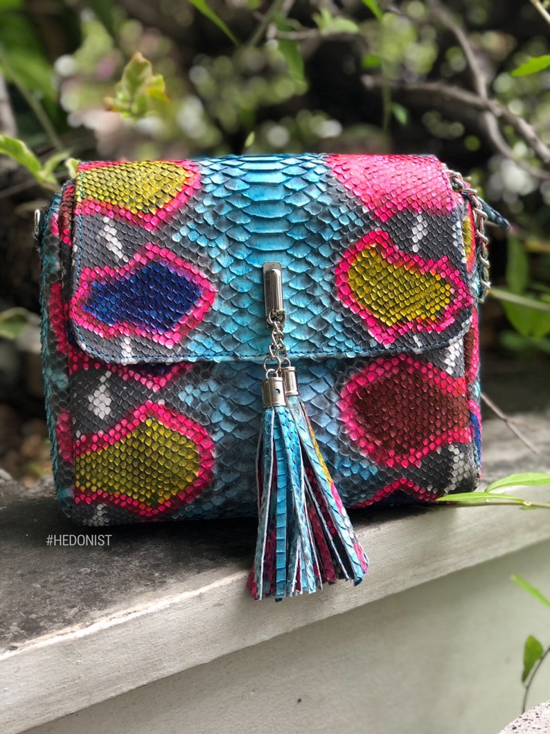 May include: A colorful crossbody bag with a snakeskin pattern in shades of blue, pink, yellow, and black. The bag features a silver chain strap and a matching tassel. The text "#HEDONIST" is visible on the bottom left.