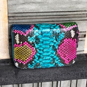 以下が含まれることがあります： ジッパー留めのカラフルなヘビ革財布。財布は本革製で、青、緑、ピンク、黄など様々な色があります。