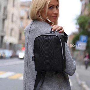 Może przedstawiać: Czarny skórzany plecak ze wzorem skóry węża. Plecak ma zamek błyskawiczny i srebrne okucia.