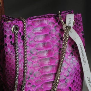 Op de afbeelding: Een klein, felroze handtasje van slangenleer met een zilveren kettingband. De tas heeft een label met de tekst "Hedonistbali" erop.