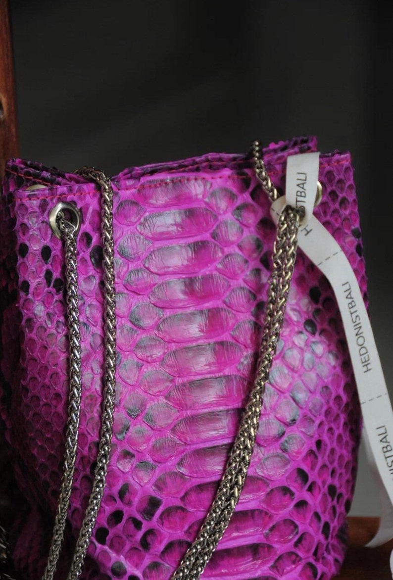 Op de afbeelding: Een klein, felroze handtasje van slangenleer met een zilveren kettingband. De tas heeft een label met de tekst "Hedonistbali" erop.