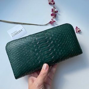 以下が含まれることがあります： ヘビ柄のダークグリーンの革財布。財布はフラップ留めになっており、小さなタグには「#ECOOUIST」と「FADE TO EDEN」の文字が書かれています。