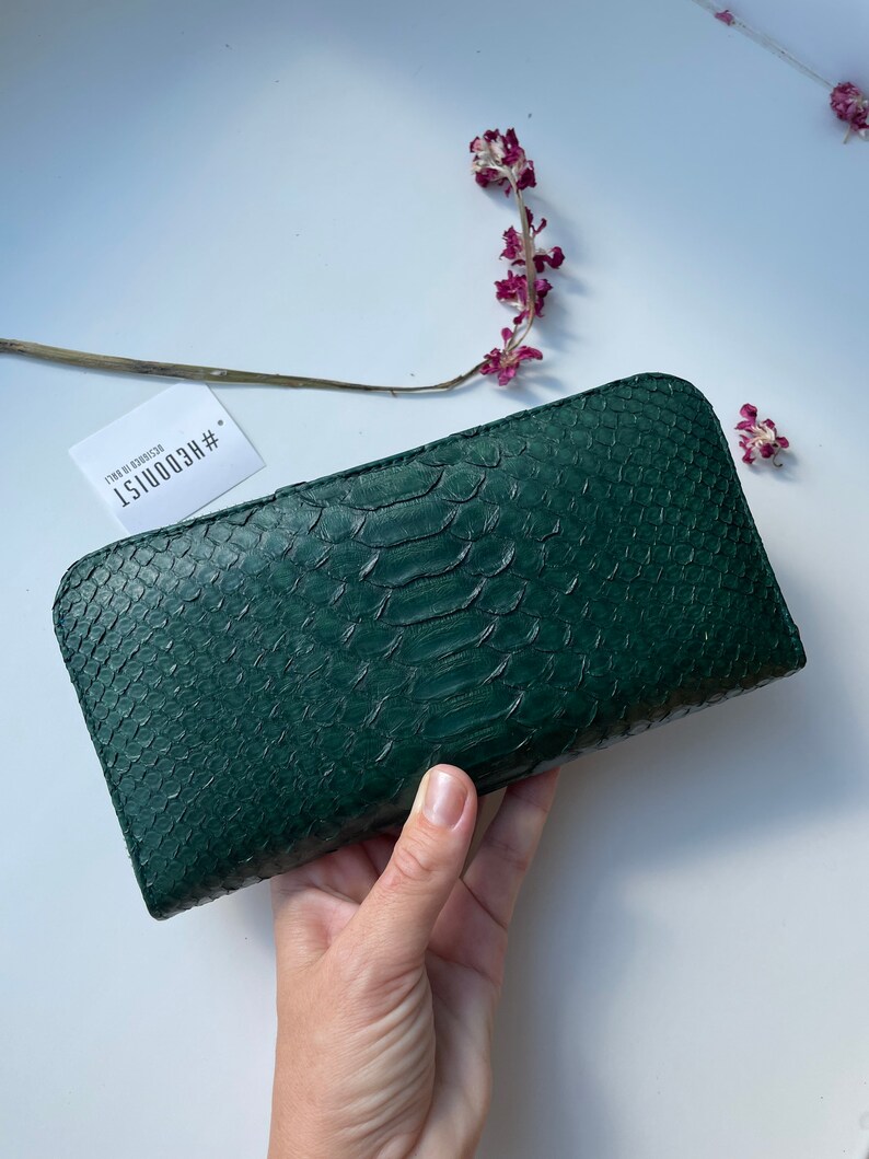 以下が含まれることがあります： ヘビ柄のダークグリーンの革財布。財布はフラップ留めになっており、小さなタグには「#ECOOUIST」と「FADE TO EDEN」の文字が書かれています。