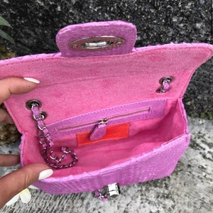 K&ouml;nnte beinhalten: Eine kleine rosa Schlangenleder-Handtasche mit einer silbernen Kettenkette und einem silbernen Verschluss. Die Tasche ist offen und das Innere ist mit rosa Stoff ausgekleidet. Im Inneren der Tasche befindet sich eine Rei&szlig;verschlusstasche.