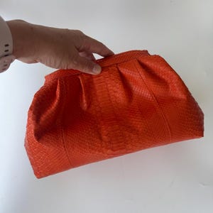 May include: A bright orange clutch bag featuring a textured snakeskin pattern. The bag has a soft, gathered, and pleated design along the top frame, creating a rounded, voluminous shape. The surface displays a consistent, repeating scale texture.