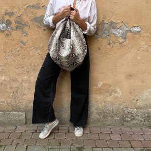 May include: A woman wearing a white button-down shirt, black pants, and sunglasses is holding a large, light brown and black snakeskin handbag.