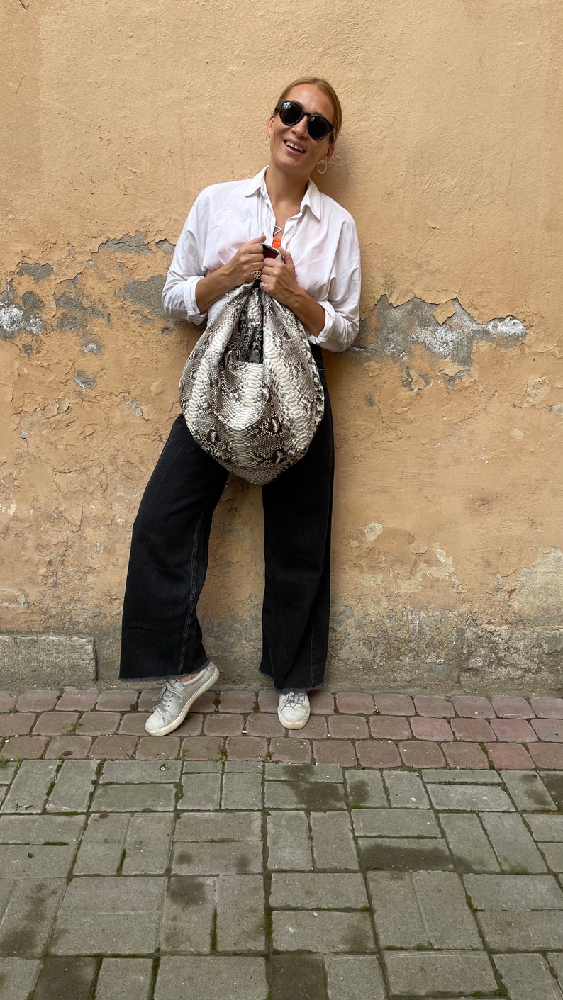 May include: A woman wearing a white button-down shirt, black pants, and sunglasses is holding a large, light brown and black snakeskin handbag.