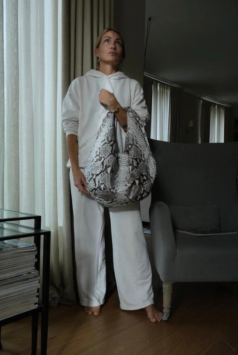May include: A woman wearing a white hoodie and matching pants is holding a large, white and gray snakeskin tote bag. The bag has a slouchy, unstructured shape.