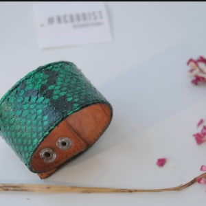 Peut inclure: Un bracelet de manchette en cuir de serpent vert avec un dos en cuir brun et deux boutons-pression argentés.