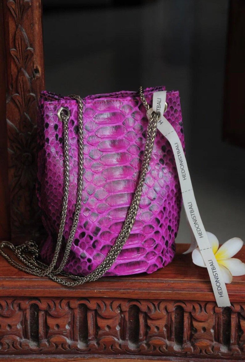 Op de afbeelding: Een klein, felpaars handtasje van slangenleer met een zilveren kettingband. De tas heeft een wit label met de tekst "Hedonistbali".