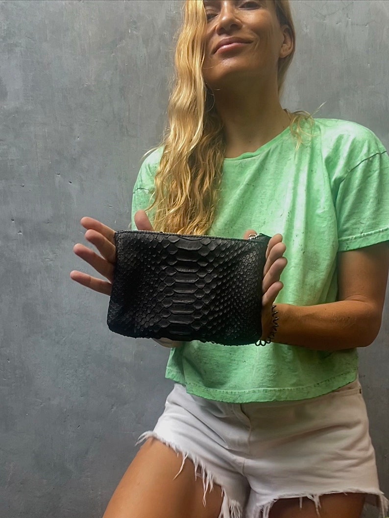 Może przedstawiać: Czarna kopert&oacute;wka ze sk&oacute;ry węża z srebrnym łańcuszkiem. Torebkę trzyma osoba ubrana w jasnozielony t-shirt i białe dżinsowe spodenki.